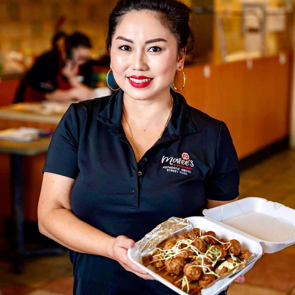 Smiling woman in a Malee's Kitchen shirt holds a takeout container of Hmong meatballs.