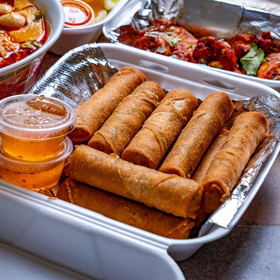 Malee's Kitchen Hmong egg rolls and dipping sauce in a takeout container, with other dishes blurred.