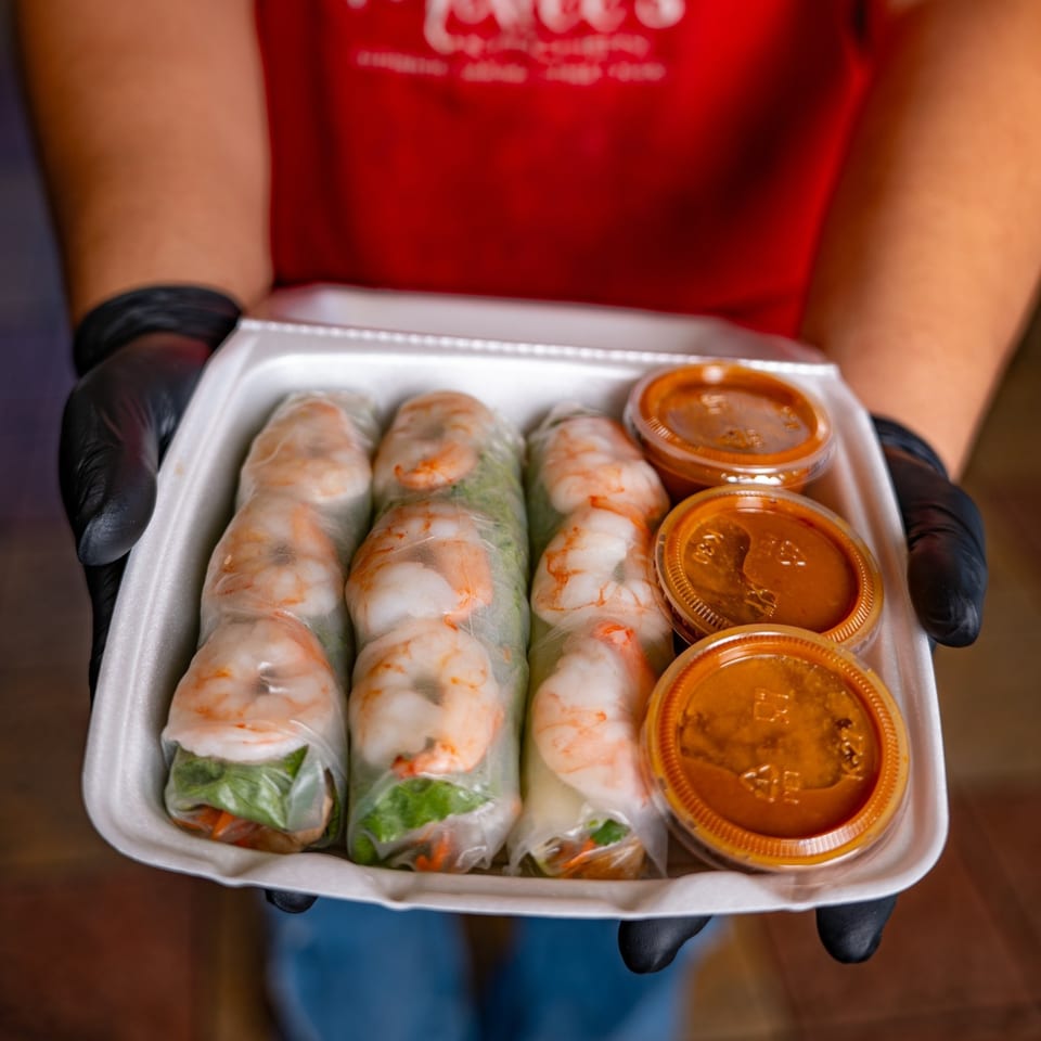 Hands in black gloves hold a takeout container with shrimp spring rolls and three dipping sauces.