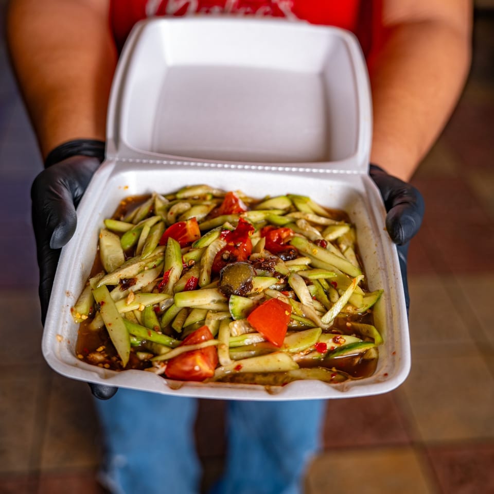 Hands in black gloves holding a white takeout container of Malee's Kitchen's Hmong cucumber salad.