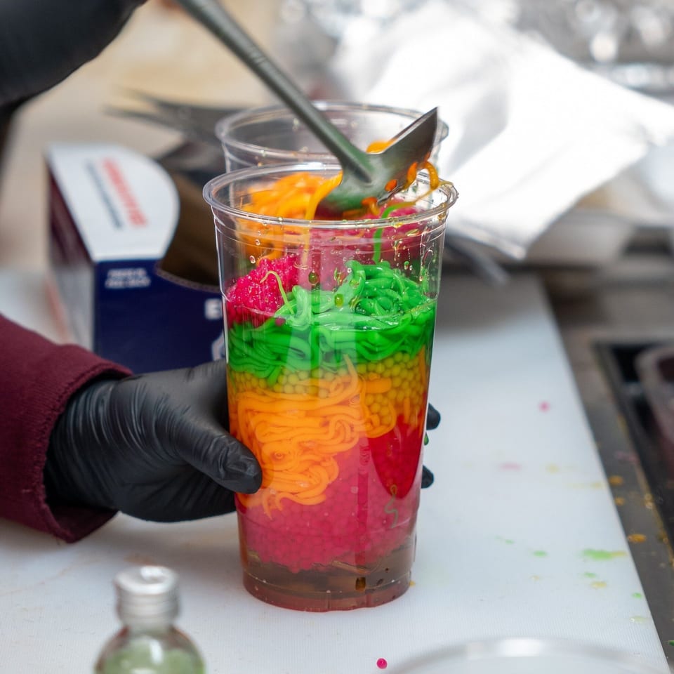 A hand in a black glove holds a clear cup filled with colorful Hmong dessert layers.