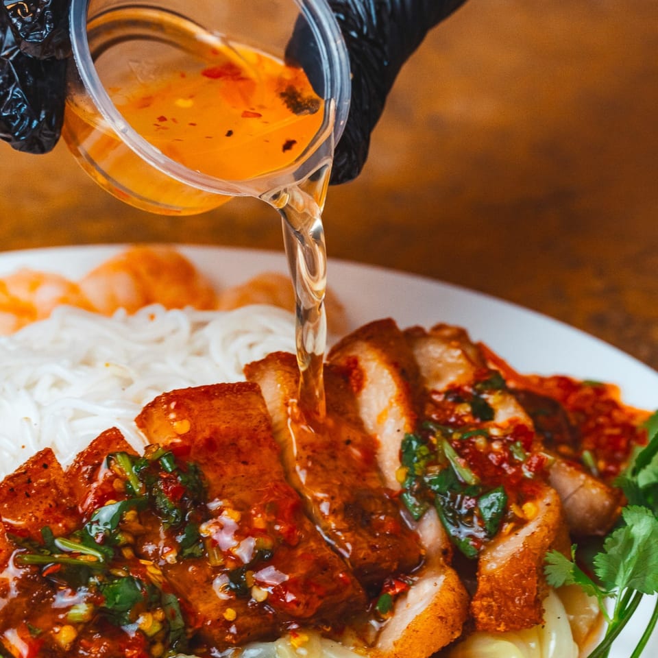A gloved hand pours sauce over crispy pork belly, noodles, and cilantro on a white plate.