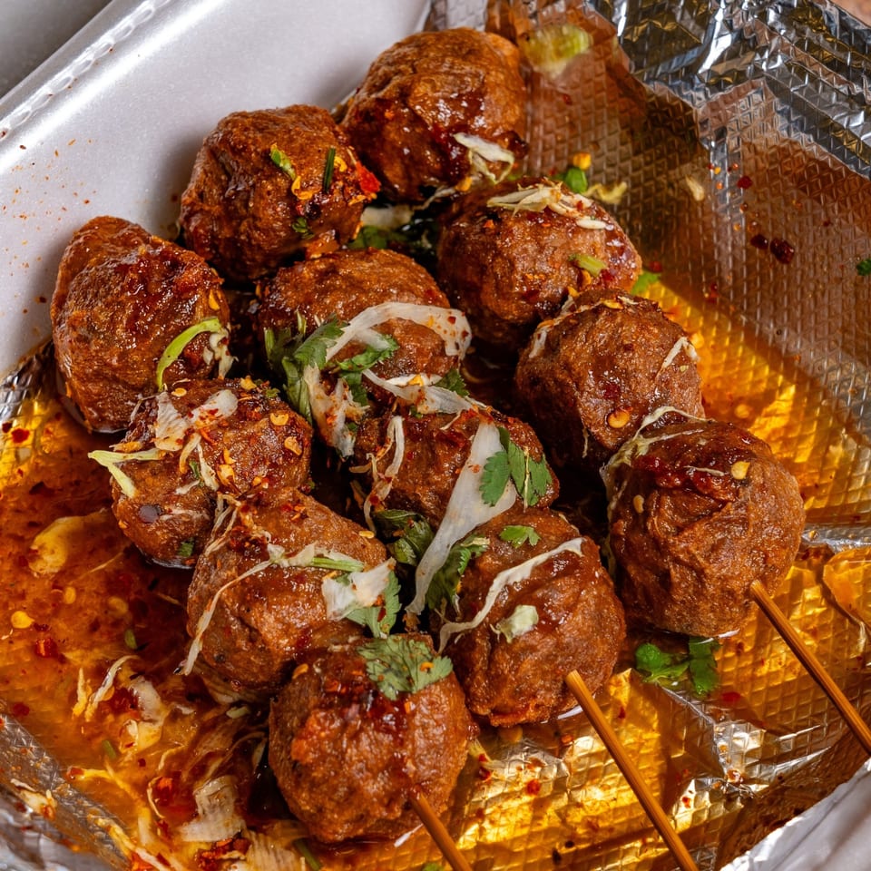 Hmong meatballs on skewers in a foil-lined container, topped with sauce, cilantro, and chili flakes.