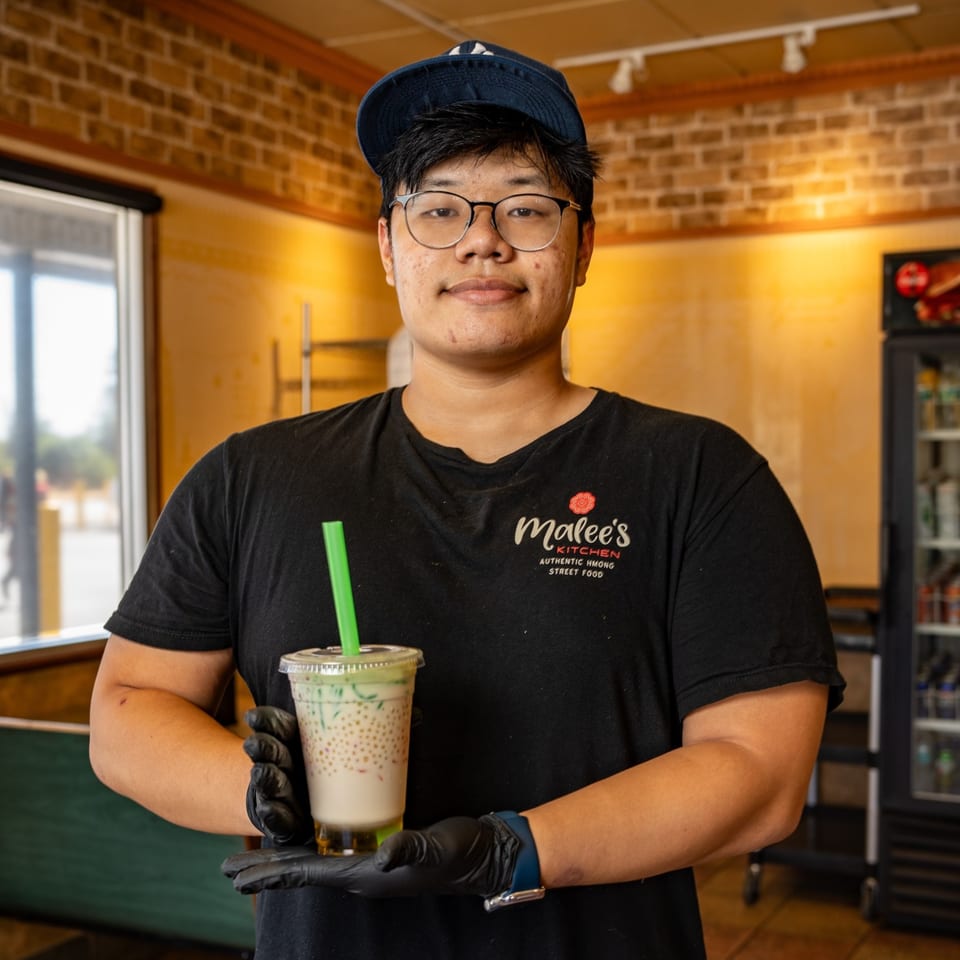 Malee's Kitchen employee in a black shirt and cap, holding a Hmong street food drink.