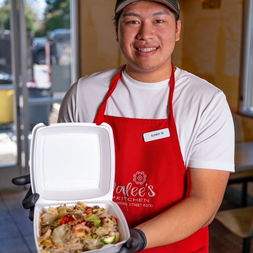 Malee's Kitchen employee, Sunny M, in a red apron, holding an open takeout container of Hmong street food.