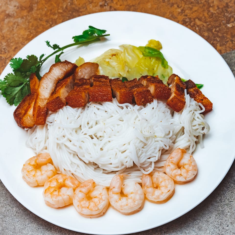 Hmong dish with crispy pork belly, rice noodles, shrimp, and cilantro on a white plate.